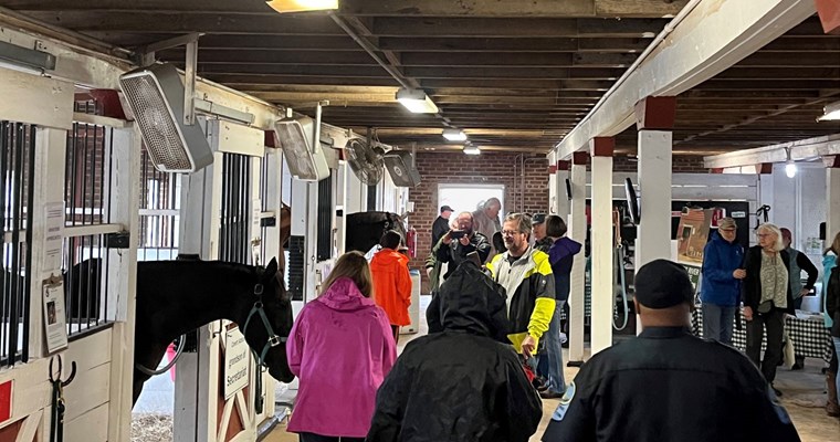 A  crowd of people gathered inside a barn with horse stalls